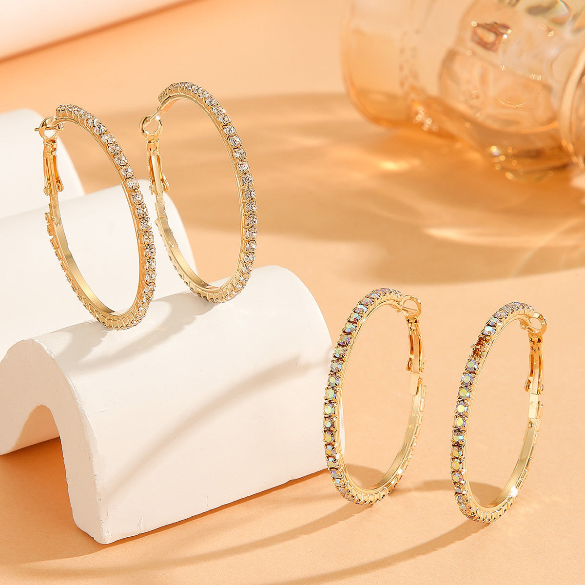 Gold hoop earrings with clear stones on a white stand against a beige background