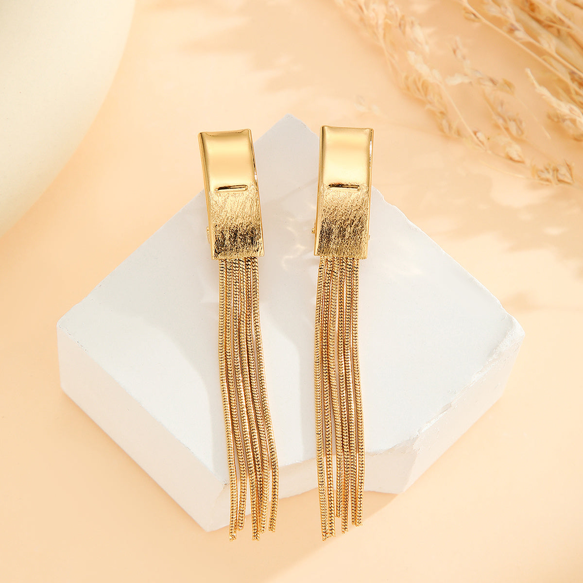 Gold fringe earrings on a white square with a beige background