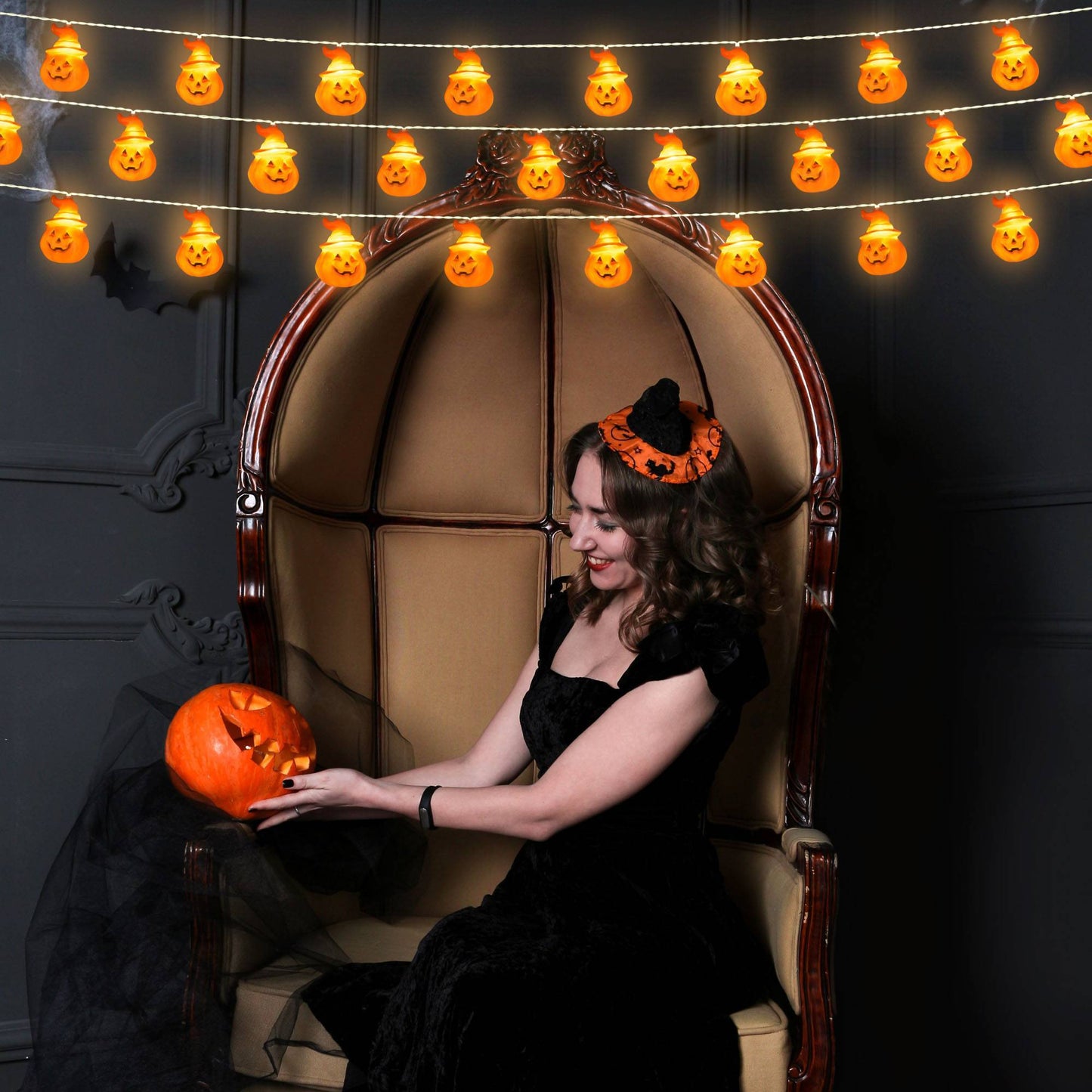 Woman in a black dress with Halloween headband sitting in a decorative chair surrounded by Halloween-themed decorations.