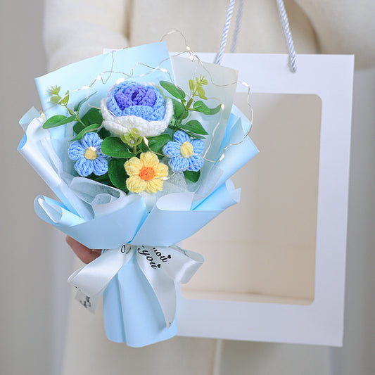 Bouquet of crocheted flowers in blue, white, and yellow with green leaves, held against a plain background.