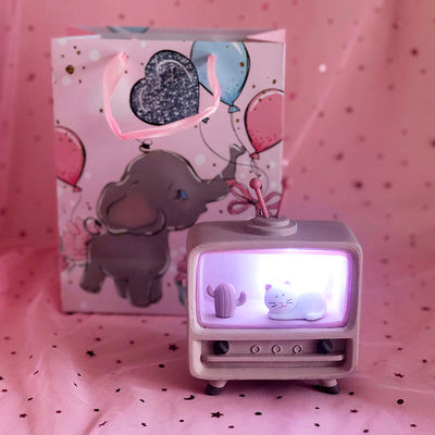 Small TV-shaped lamp with a cat design on a pink surface with a card featuring an elephant and balloons in the background.