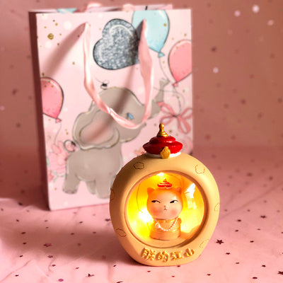 Decorative lamp with a cartoon character inside, placed on a pink surface with a matching gift box in the background.