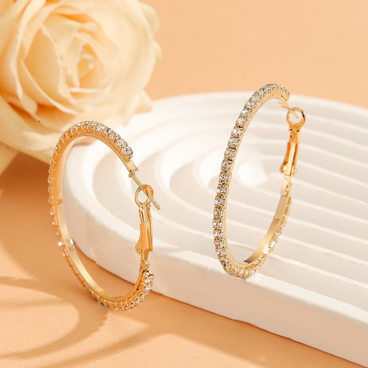 Gold hoop earrings with clear stones on a white surface with a beige flower in the background