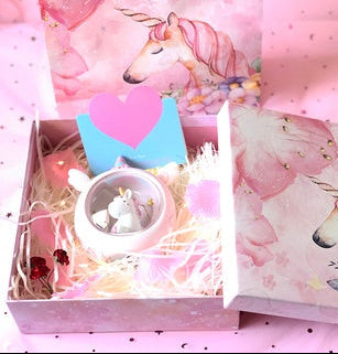 Unicorn-themed gift set with a box, card, and small figurine on a pink background.