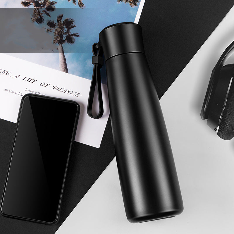 Black water bottle on a white surface with a smartphone and magazine underneath